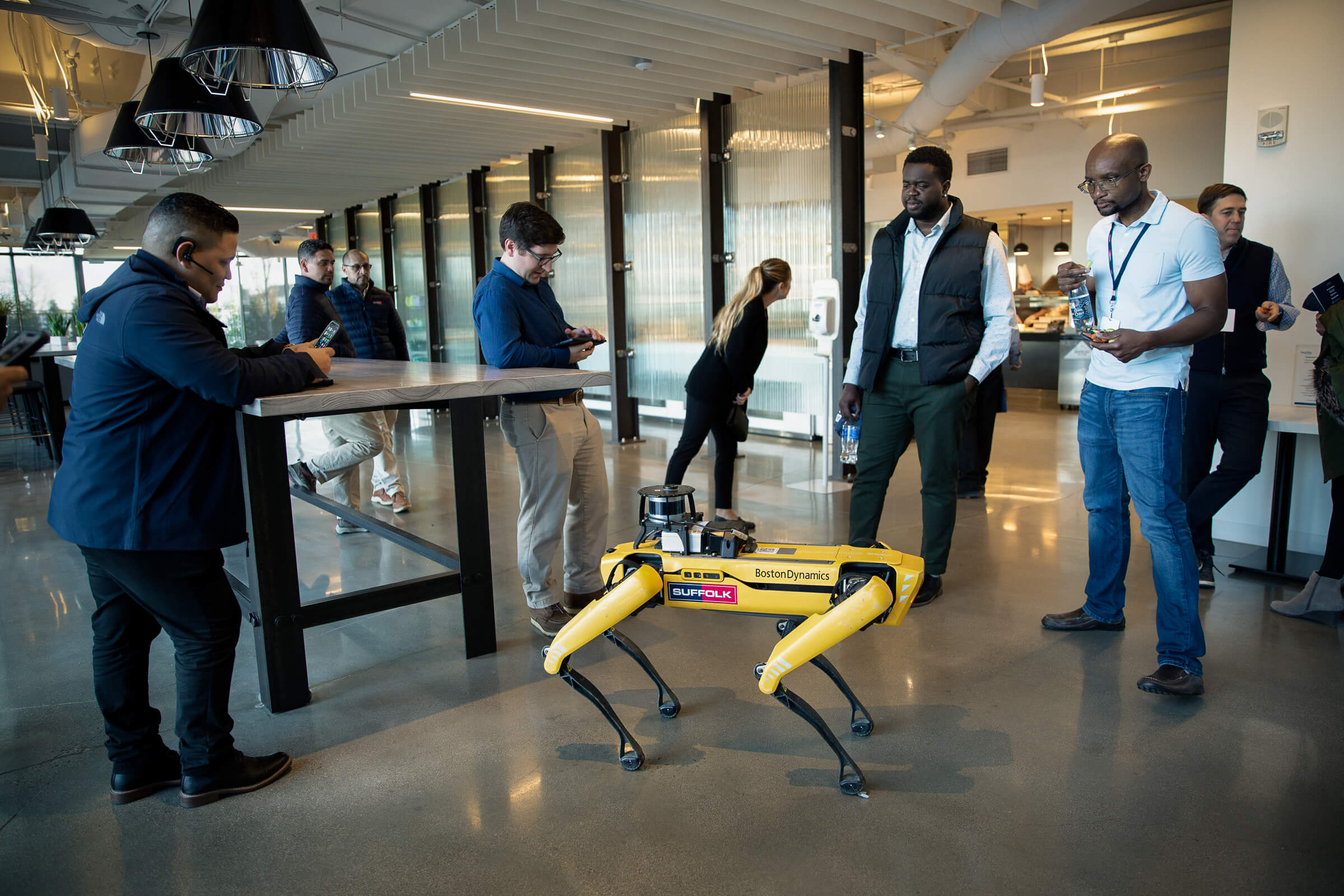 Group of professional office workers looking at a yellow robotics machine in a brightly lit office space.